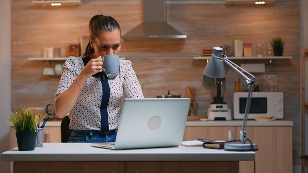 Business woman working from home late at night writing on laptop and drinking coffee. Busy focused employee using modern technology network wireless doing overtime for job reading typing, searching
