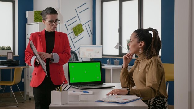 Business women using laptop with green screen and talking about work. Colleagues with mockup template and isolated background on chroma key computer display. Mock-up copy space screen