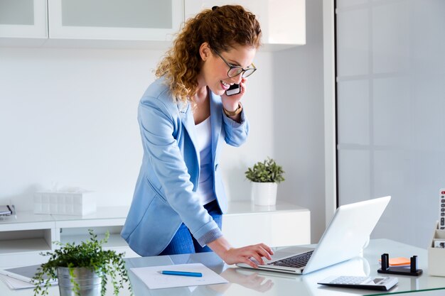 Деловая молодая женщина, говорить на мобильном телефоне, используя ее ноутбук в офисе.