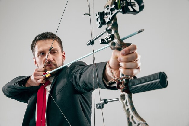 Businessman aiming at target with bow and arrow, isolated on gray studio wall