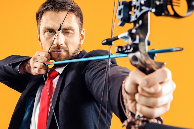 Free photo businessman aiming at target with bow and arrow, isolated on yellow studio background. the business, goal, challenge, competition, achievement concept