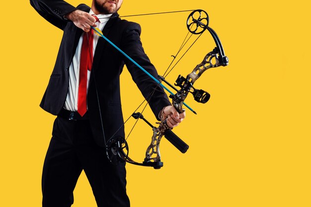 Businessman aiming at target with bow and arrow, isolated on yellow studio wall