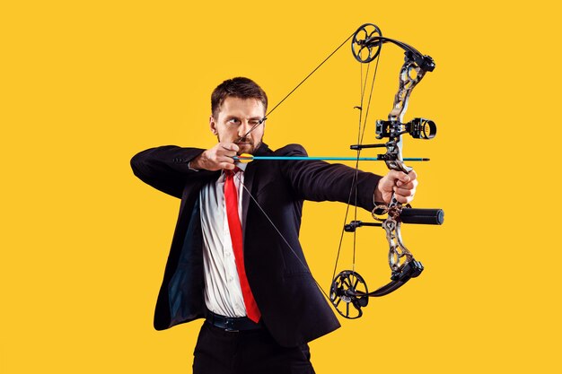 Businessman aiming at target with bow and arrow, isolated on yellow studio wall