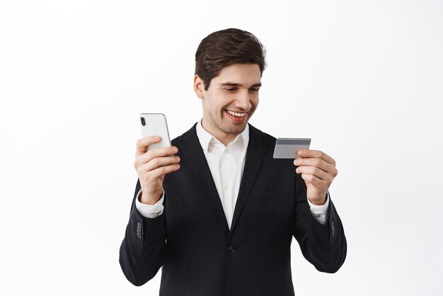 검은 양복을 입은 사업가는 스마트폰 앱에서 온라인 쇼핑으로 온라인 결제를 하는 동안 신용 카드를 보고 비접촉식 흰색 배경을 주문합니다.