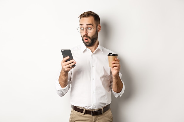 Бизнесмен пьет кофе и смотрит на сообщение на мобильном телефоне с удивлением, стоя в изумлении