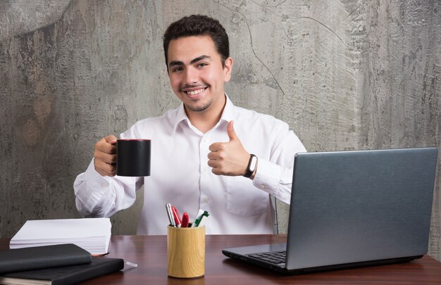 Businessman holding cup of tea and showing thumbs up at the office desk.