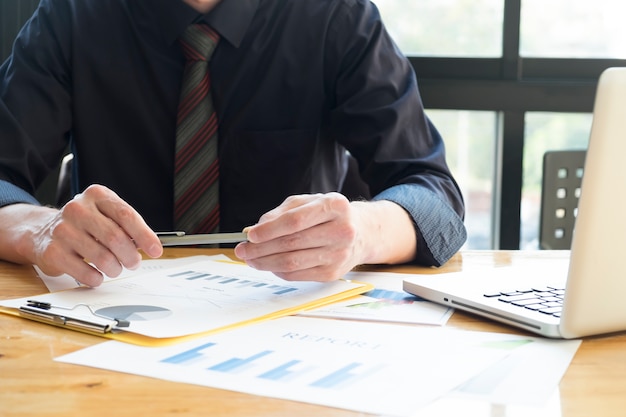 Businessman holding pen analyzing investment charts with laptop.