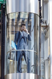 Бизнесмен в современном стеклянном лифте разговаривает по телефону и держит документы