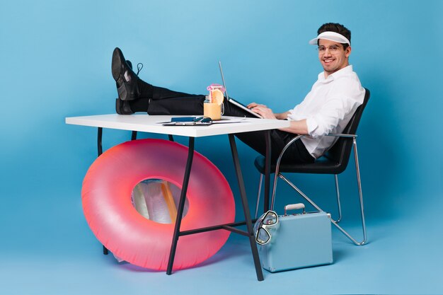 Businessman packed suitcase on vacation and happily working. Guy in office clothes holds laptop and looks into camera on blue space with inflatable circle.