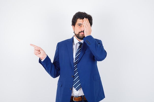 Businessman pointing left with index fingers and covering eye in formal suit and looking serious , front view.