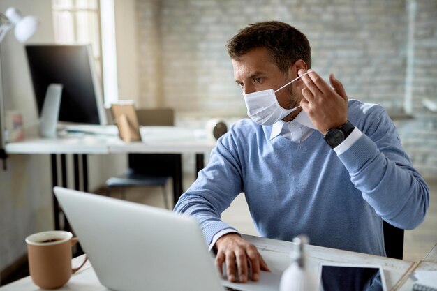 Бизнесмен надевает защитную маску во время работы на ноутбуке в офисе