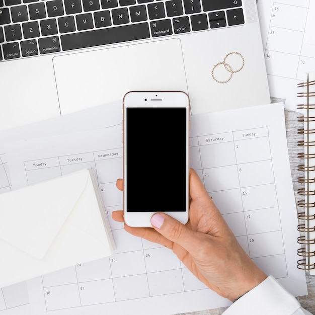 Businessman's hand holding smartphone over the calendar with envelope; laptop and wedding rings