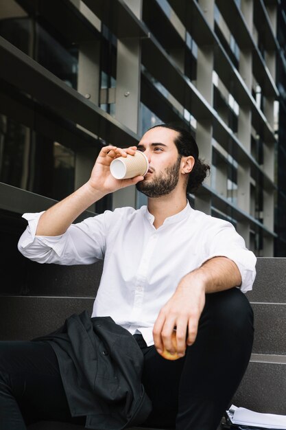 Бизнесмен сидит возле офисного здания с яблоком в руке и пьет кофе