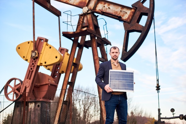 Businessman standing on an oilfield holding mini solar module next to an oil rig