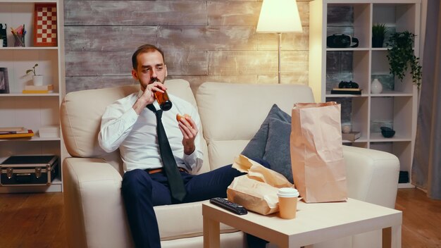 Businessman in suit sitting on couch eating a burger, drinking beer and watching tv.