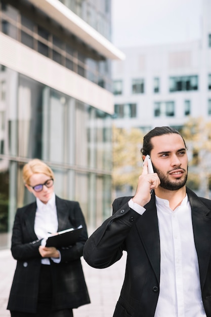 Businessman talking on mobile phone and businesswoman writing on clipboard in the background