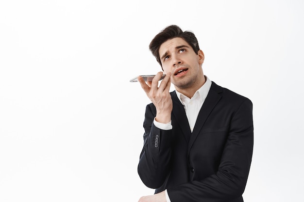 Businessman talking on speakerphone, look up thoughtful and record voice on smartphone, using translator app on cellphone, standing over white wall