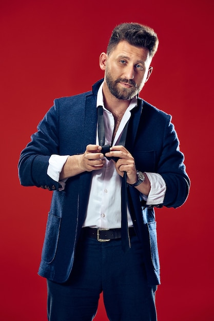 Businessman tying his tie at studio. Smiling business man standing isolated on red studio background. Beautiful male half-length portrait