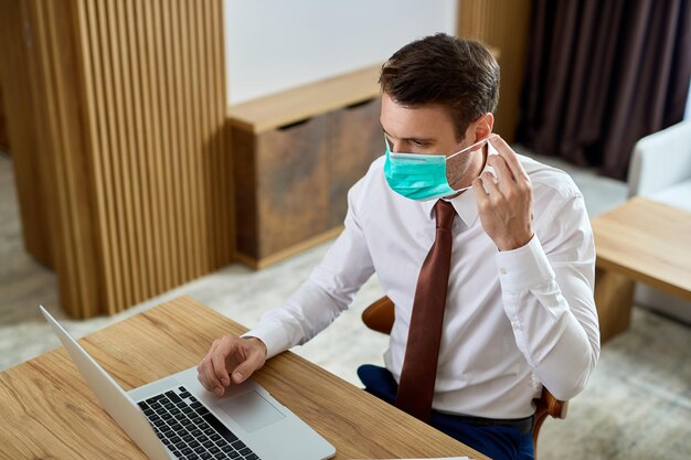 Businessman using laptop and putting on protective face mask while working in a hotel room