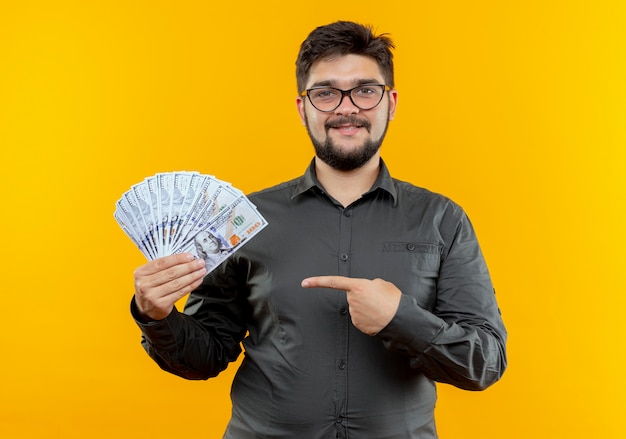 Businessman wearing glasses holding and points at cash isolated on yellow