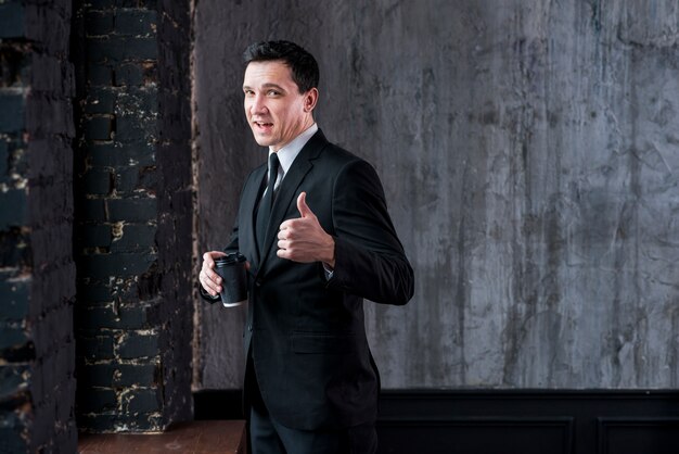 Businessman with coffee cup showing thumb up and looking at camera