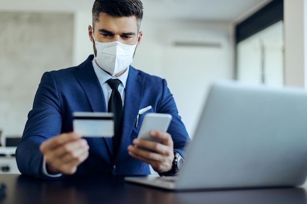 Uomo d'affari con maschera facciale ebanking con carta di credito e smartphone in ufficio