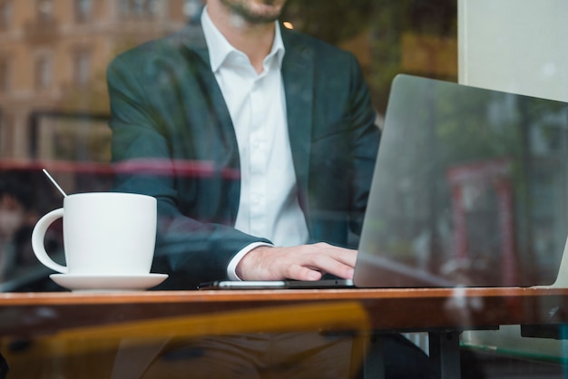 Businessman working on laptop in cafe seen through glass