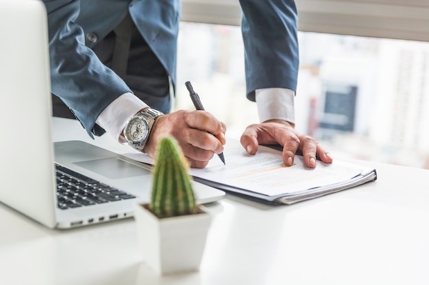 Businessman writing on document with pen on desk with laptop