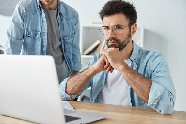 Businessmen sit at office desk together, work and communicate, concentrated into laptop computer