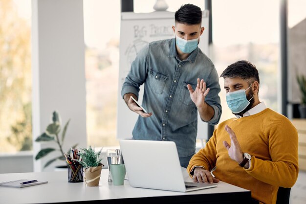 Бизнесмены в масках машут во время видеозвонка через ноутбук в офисе