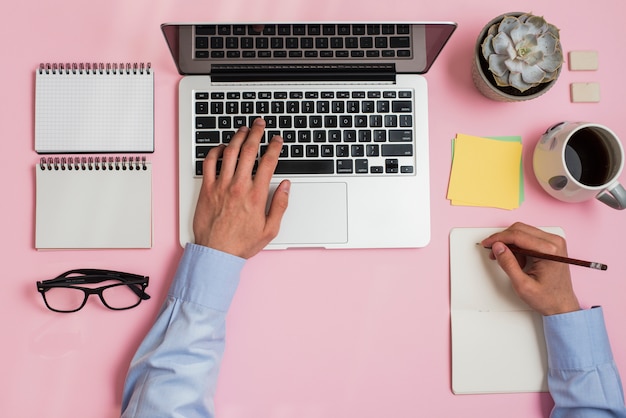 A businessperson writing on notepad typing on laptop over the office desk