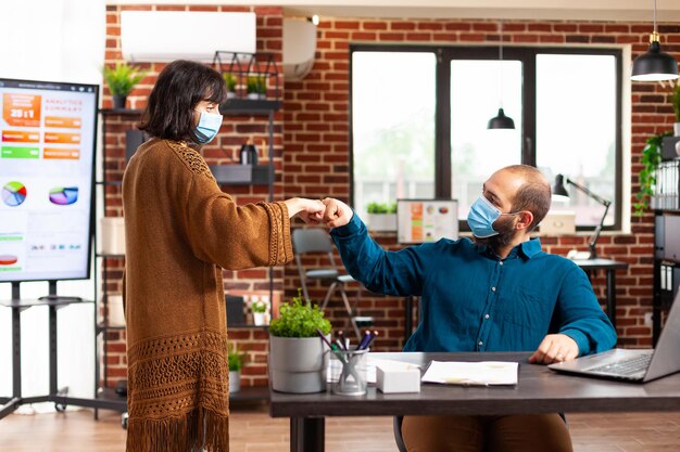 Businessteam with medical protective face mask against coronavirus greeting giving first bump in startup office. Manager man sitting at desk brainstorming ideas for business project typing strategy