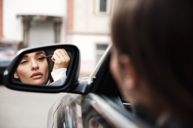 Businesswoman driver looking at rear-view mirror while sitting in car and applying mascara