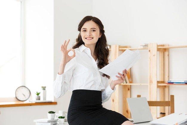 Businesswoman in formal wear holding documents and showing ok sign at modern office copy space