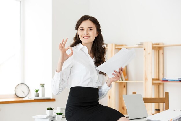 Деловая женщина в формальной одежде держит документы и показывает знак "ок" в современном офисе