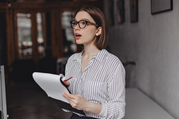 Businesswoman in glasses and white classic shirt takes notes in papers and poses in office.