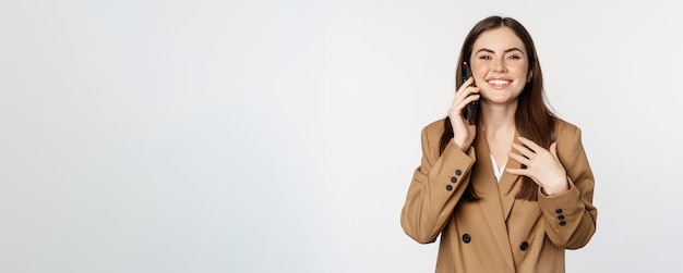Businesswoman having mobile call conversation on smartphone talking with client standing over white background in brown suit