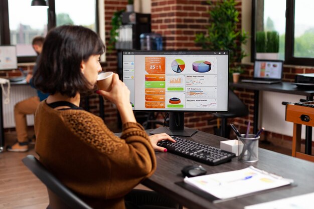 Businesswoman holding cup of coffee analyzing financial graphs typing marketing strategy while working at company project in startup office. Executive manager researching business solution