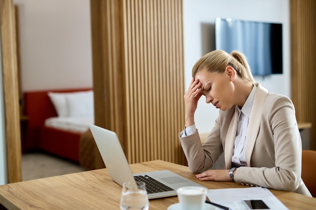 Деловая женщина держится за голову от боли и чувствует себя измотанной после работы за компьютером в гостиничном номере