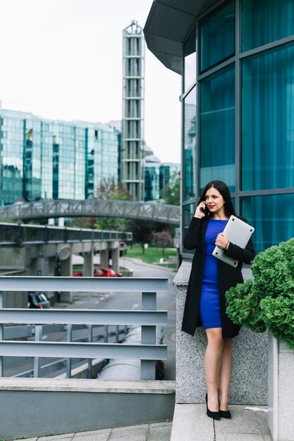 Businesswoman holding laptop and talking on mobile phone outside office building