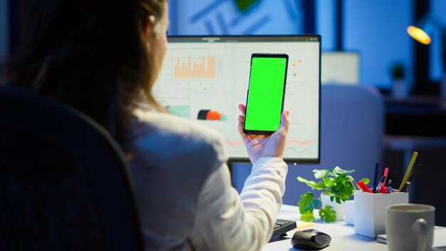 Businesswoman looking at green screen monitor of smartphone sitting at desk in business office late at night. Freelancer watching desktop monitor display with green mockup, chroma key working overtime