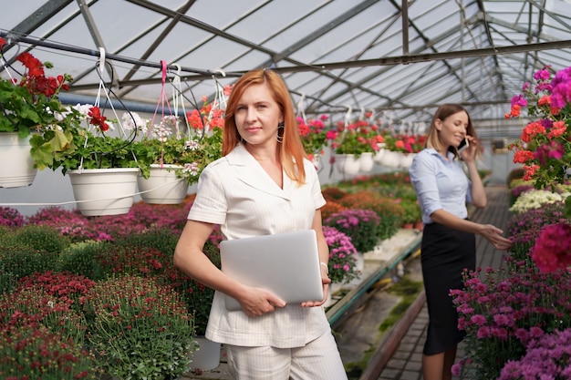 Businesswoman posing with a laptop while her partner discussing on the phone a proposal in a green house with flowers.