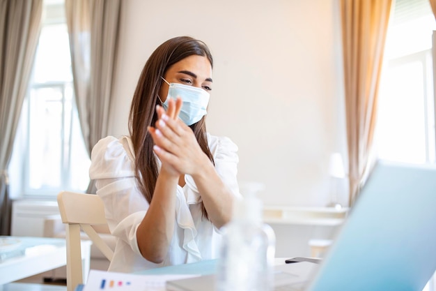 Businesswoman putting on protective mask working on laptop in the office Focused company employee worker in protective facial mask working on laptop at home keeping covid 19 virus quarantine measures