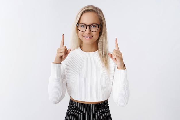 Businesswoman sharing secrets of success pointing up with index fingers and smiling with encouraging assuring grin posing in glasses and stylish cropped sweater against white background.