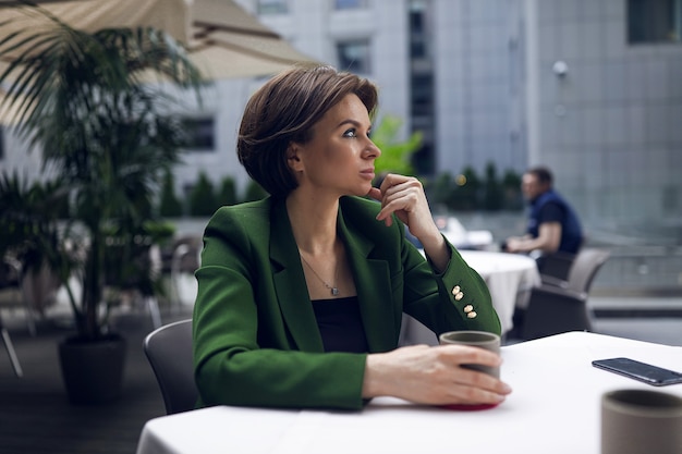 Businesswoman sitting in cafe and having some rest after all meetings and interviews. Green stylish jacket and black blouse, short haircut, nude makeup. Cup of hot coffee
