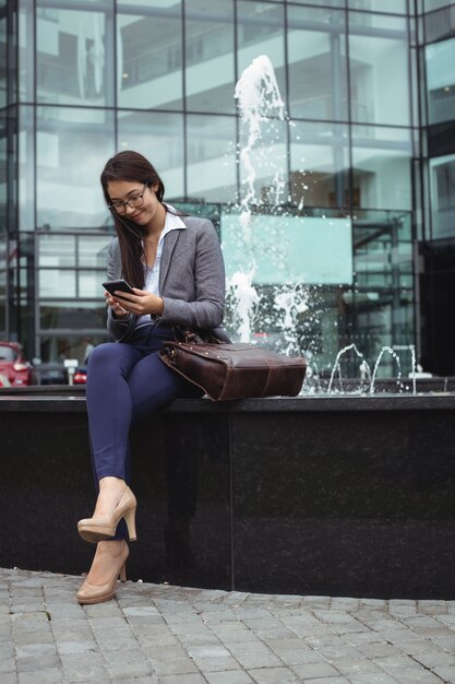 Businesswoman sitting near fountain and using mobile phone