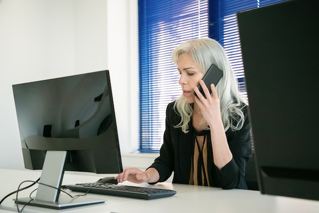 Businesswoman sitting in office and talking on phone. Grey-haired confident employee typing on computer keyboard and discussing work with client via smartphone. Business and communication concept