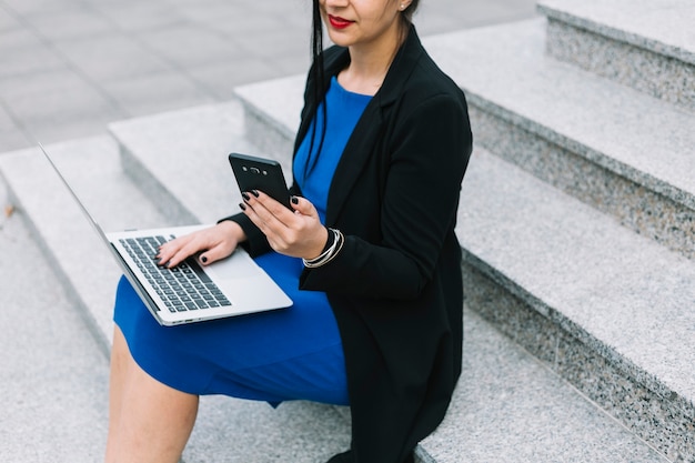 Businesswoman sitting on staircase using laptop and smartphone