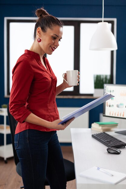 Businesswoman standing up next to desk holding clipboard and cup. Entrepreneur in startup office looking at business results and drinking coffee. Employee in red shirt working casually.