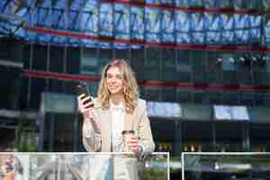 Foto gratuita imprenditrice in giacca e cravatta in piedi con lo smartphone in strada a bere caffè utilizzando la fronte dell'app del telefono cellulare